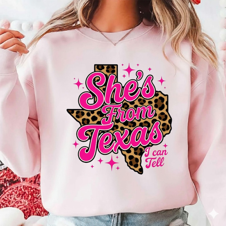 May include: A light pink sweatshirt with the text "She's From Texas I can Tell" in a pink and black design. The design includes a leopard print Texas outline and pink star accents.
