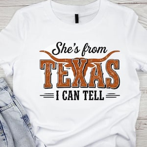 May include: White t-shirt with the text "She's from TEXAS I CAN TELL" in a distressed, brown and black design. The word "TEXAS" is in large, bold letters with a longhorn graphic above it.