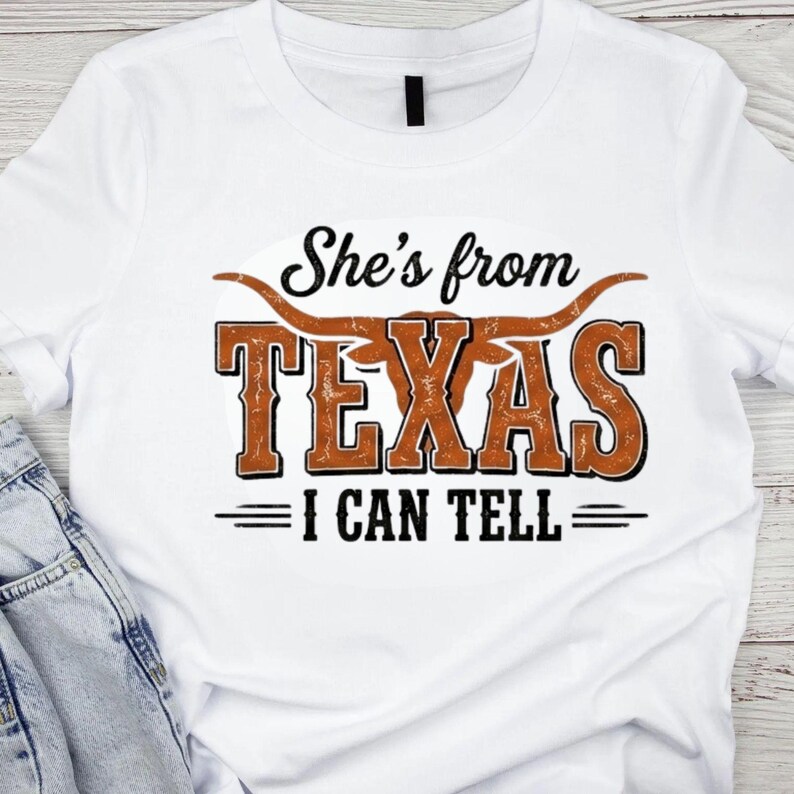 May include: White t-shirt with the text "She's from TEXAS I CAN TELL" in a distressed, brown and black design. The word "TEXAS" is in large, bold letters with a longhorn graphic above it.