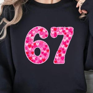 May include: Black sweatshirt featuring the number "67" in a pink heart pattern. The numbers are outlined in a darker pink, with a variety of pink and red hearts filling the numbers. The sweatshirt is a solid black color.