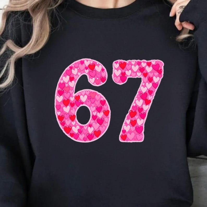 May include: Black sweatshirt featuring the number "67" in a pink heart pattern. The numbers are outlined in a darker pink, with a variety of pink and red hearts filling the numbers. The sweatshirt is a solid black color.