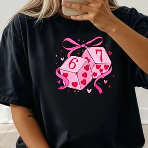 May include: Black t-shirt featuring a graphic of two pink dice with the numbers 6 and 7. The dice are decorated with red hearts and tied together with a pink ribbon and bow. Small hearts and stars are scattered around the dice.
