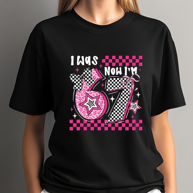 May include: Black t-shirt with a graphic design featuring the text "I WAS 67 NOW I'M" in white. The design includes a pink number 6 with a leopard print pattern and a checkered bow, and a checkered number 7. Pink and white stars and a checkered border complete the design.