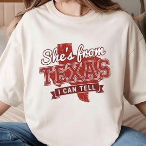 May include: Cream-colored t-shirt with a distressed red graphic that reads "She's from TEXAS I CAN TELL." The design features the outline of the state of Texas. The shirt is a casual, unisex style.