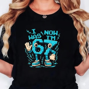 May include: Black t-shirt with a cartoon graphic of the numbers 67 as animated characters. The text "I WAS NOW I'M" is above the numbers. The numbers are light blue with black outlines and lightning bolt accents.