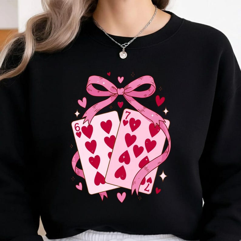 May include: Black sweatshirt featuring a pink bow and two playing cards with red hearts. The cards display the numbers six and seven. The design includes small hearts and starbursts, creating a romantic theme.