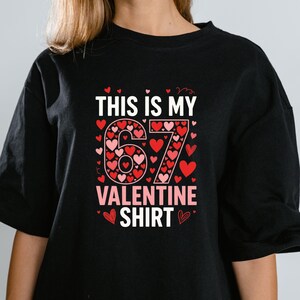 May include: Black t-shirt with the text "THIS IS MY 67 VALENTINE SHIRT". The number 67 is designed with red and pink hearts. The words "VALENTINE SHIRT" are in pink. Small red hearts are scattered around the text.