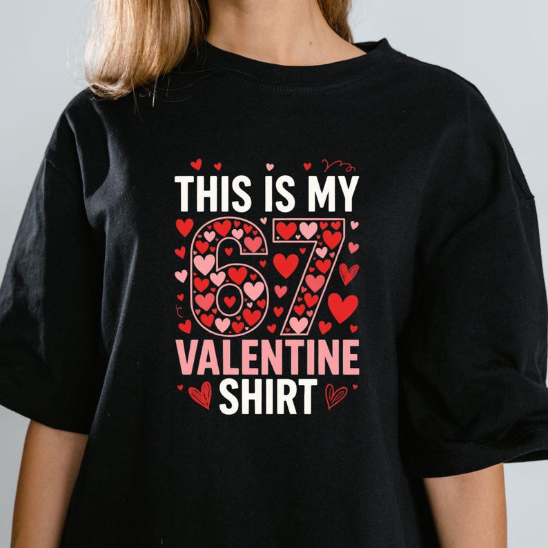 May include: Black t-shirt with the text "THIS IS MY 67 VALENTINE SHIRT". The number 67 is designed with red and pink hearts. The words "VALENTINE SHIRT" are in pink. Small red hearts are scattered around the text.