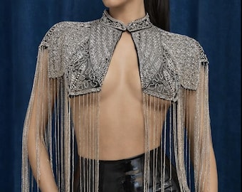 May include: A cropped bolero jacket featuring intricate silver beadwork and embroidery on a dark base. The garment has a high collar, short structured shoulders, and long, flowing silver fringe that extends from the hem down to the waist.
