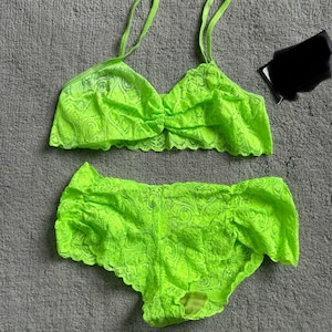 May include: A neon green lace lingerie set, including a bralette with thin straps and a matching pair of shorts. The set is made of a delicate, see-through lace fabric. The bralette has a gathered center detail. The shorts have a ruffled edge.