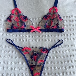 May include: A two-piece lingerie set consisting of a bralette and a thong. Both pieces are made of sheer nude mesh fabric featuring dark blue and neon pink floral embroidery. The bralette has a neon pink bow at the center and dark blue straps.