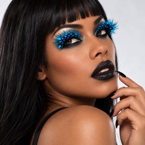 May include: Dramatic false eyelashes featuring bright blue, spiky, feathered tips with black bases. The lashes are applied over dark, smoky eyeshadow. The look is complemented by matte black lipstick and long, pointed black fingernails.