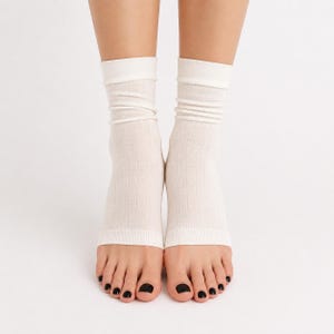 May include: A pair of off-white, ribbed, open-toe socks that cover the ankles and lower shins. The socks feature a fine vertical knit texture, a reinforced cuff at the top, and a straight, hemmed edge across the mid-foot, leaving the toes exposed.