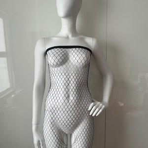 May include: A white mannequin wearing a black fishnet body stocking. The body stocking is a strapless design with a diamond pattern. The mannequin is positioned against a white wall.