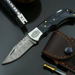 May include: A folding knife with a patterned blade and a black handle with gold accents. The blade has a unique design, and the handle is engraved with the name "DANIEL". A brown leather sheath is also included.