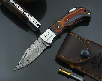 May include: A folding pocket knife with a patterned blade and a wooden handle. The knife has the name "JACK" and the word "Groomsman" engraved on the blade. A brown leather sheath is also included.