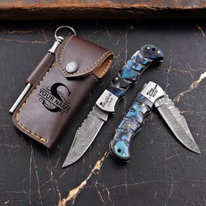 Puede incluir: Un juego de tres cuchillos con una funda de cuero marrón. Dos cuchillos plegables tienen mangos con diseños azules y negros y hojas plateadas. La funda está grabada con el texto "YOUR NAME" y la letra "S". Se incluye una varilla de afilado.