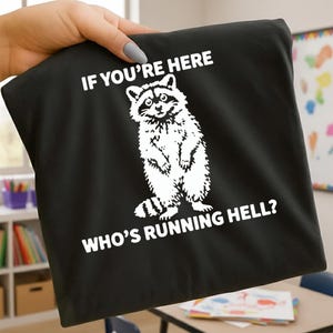 Könnte beinhalten: Schwarzes T-Shirt mit einer weißen Grafik eines Waschbären und dem Text "IF YOU'RE HERE" über dem Waschbären und "WHO'S RUNNING HELL?" darunter. Der Waschbär steht aufrecht.