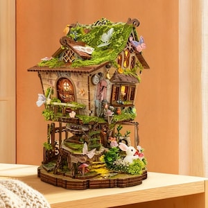 Puede incluir: Una detallada casa de hadas de varios niveles con un tejado cubierto de musgo, pequeñas ventanas y un exterior de madera. La casa está adornada con figuras en miniatura, mariposas y flores. Se encuentra sobre una estantería de madera con libros y una manta.