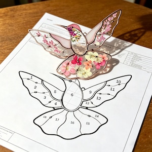 Puede incluir: Un colibrí de vidriera con bordes color cobre, relleno de flores secas en tonos rosa, morado y amarillo. Las alas son translúcidas. Debajo hay un diagrama en blanco y negro del colibrí.