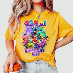 Pode incluir: Camiseta amarela com um gr&aacute;fico colorido com o texto "MOMMY OF THE BIRTHDAY BOY". O design inclui v&aacute;rios personagens de desenhos animados. A camiseta &eacute; feita de um material macio.