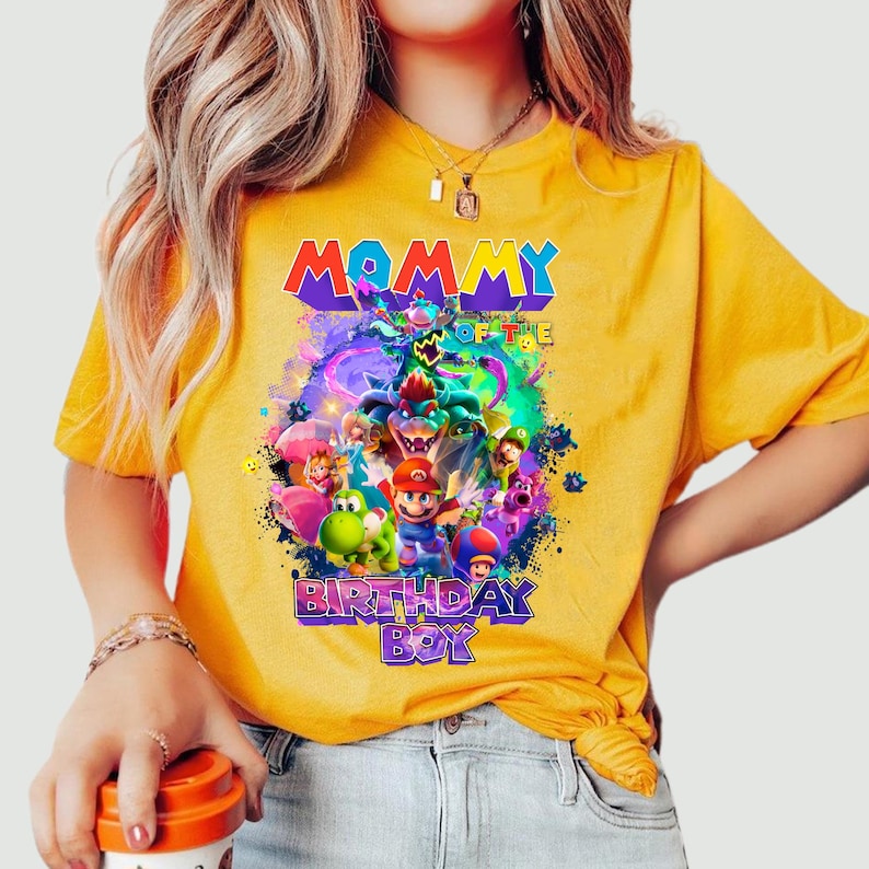 Pode incluir: Camiseta amarela com um gr&aacute;fico colorido com o texto "MOMMY OF THE BIRTHDAY BOY". O design inclui v&aacute;rios personagens de desenhos animados. A camiseta &eacute; feita de um material macio.