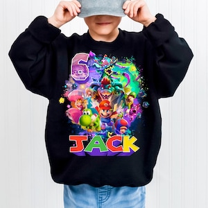Pode incluir: Camisola preta com um gr&aacute;fico colorido de personagens de videojogos, o n&uacute;mero 6 e o nome "JACK" em letras 3D. O design apresenta personagens de uma popular s&eacute;rie de videojogos.