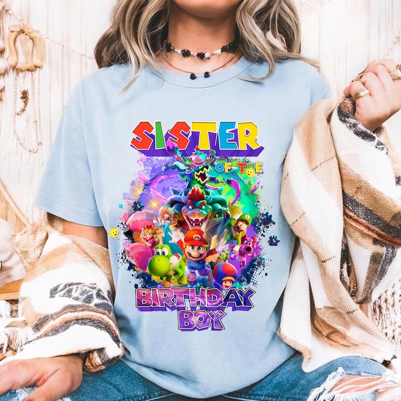 Pode incluir: Camiseta azul clara com um gr&aacute;fico colorido. O gr&aacute;fico inclui o texto "SISTER OF THE BIRTHDAY BOY" e personagens de desenhos animados em um design vibrante. A camiseta &eacute; feita de um material macio.