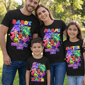 Pode incluir: Quatro camisetas pretas com gr&aacute;ficos coloridos e texto. As camisetas dizem "Daddy", "Mommy", "Sister" e "Jack". A camiseta "Jack" tamb&eacute;m tem o n&uacute;mero "6". Os gr&aacute;ficos apresentam personagens de videogame. A fam&iacute;lia est&aacute; sorrindo.