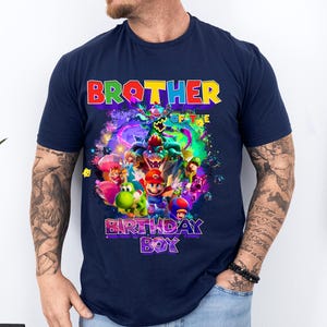 Pode incluir: Camiseta azul marinho com um gr&aacute;fico colorido com personagens de desenhos animados e o texto "BROTHER OF THE BIRTHDAY BOY". O design inclui personagens em vermelho, verde, azul e amarelo.