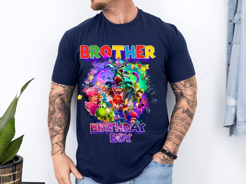 Pode incluir: Camiseta azul marinho com um gr&aacute;fico colorido com personagens de desenhos animados e o texto "BROTHER OF THE BIRTHDAY BOY". O design inclui personagens em vermelho, verde, azul e amarelo.