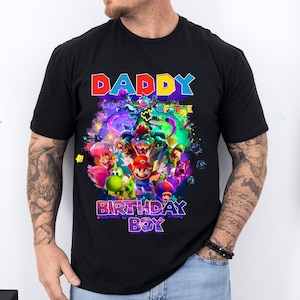Pode incluir: Camiseta preta com um gr&aacute;fico colorido de personagens de videogame, incluindo Mario e Yoshi. O texto "DADDY OF THE BIRTHDAY BOY" &eacute; exibido em cores vibrantes. A camisa &eacute; de manga curta e parece ser de algod&atilde;o.