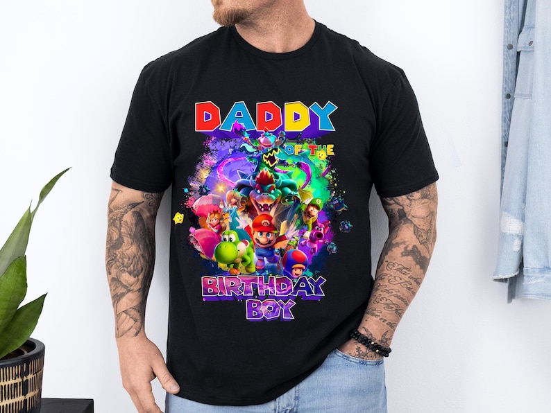 Pode incluir: Camiseta preta com um gr&aacute;fico colorido de personagens de videogame, incluindo Mario e Yoshi. O texto "DADDY OF THE BIRTHDAY BOY" &eacute; exibido em cores vibrantes. A camisa &eacute; de manga curta e parece ser de algod&atilde;o.