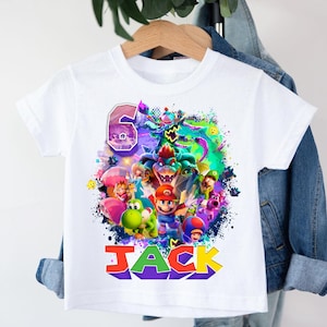 Pode incluir: Camiseta branca com um gr&aacute;fico colorido de personagens de videojogos, o n&uacute;mero 6 e o nome "JACK" em fonte 3D. O design inclui personagens de uma popular franquia de videojogos. A camiseta &eacute; exibida em um cabide.