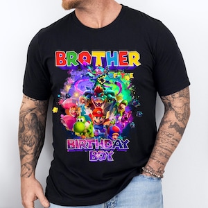 Pode incluir: Camiseta preta com um gr&aacute;fico colorido com personagens de videogame. O texto "BROTHER OF THE BIRTHDAY BOY" &eacute; exibido em cores vibrantes. O design inclui figuras de desenhos animados e um fundo din&acirc;mico e multicolorido.