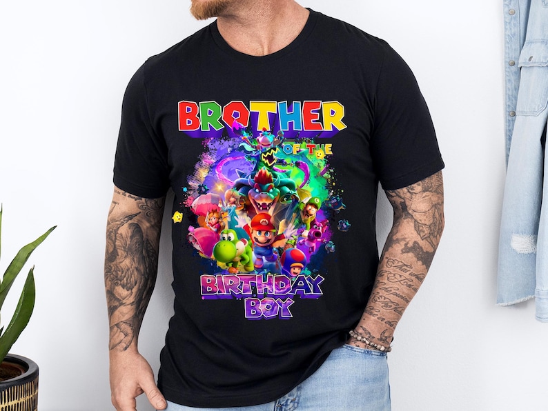 Pode incluir: Camiseta preta com um gr&aacute;fico colorido com personagens de videogame. O texto "BROTHER OF THE BIRTHDAY BOY" &eacute; exibido em cores vibrantes. O design inclui figuras de desenhos animados e um fundo din&acirc;mico e multicolorido.