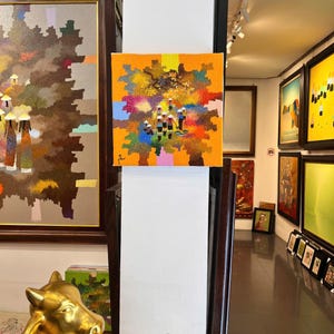 Può includere: Una piccola opera d'arte quadrata con sfondo arancione e una colorata rappresentazione astratta di figure con cappelli conici. L'opera è esposta in una galleria, circondata da altri dipinti e sculture incorniciate.