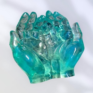 May include: A translucent teal-colored sculpture of two cupped hands. The hands are detailed with visible fingers and a textured surface, creating a sense of depth. The sculpture has a glossy finish and is set against a white background.