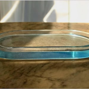 May include: An oval glass tray with a light blue edge. The tray is on a brown, marble-effect surface, with sunlight reflecting off the glass. The tray is empty and appears to be a decorative item. The tray's dimensions are not visible.