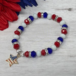 Pu&ograve; includere: Un braccialetto patriottico con perline rosse, bianche e blu, intervallate da distanziatori argentati. Un ciondolo a forma di stella dorata con un design a bandiera americana pende dal braccialetto. Il braccialetto &egrave; esposto su una superficie in legno chiaro.
