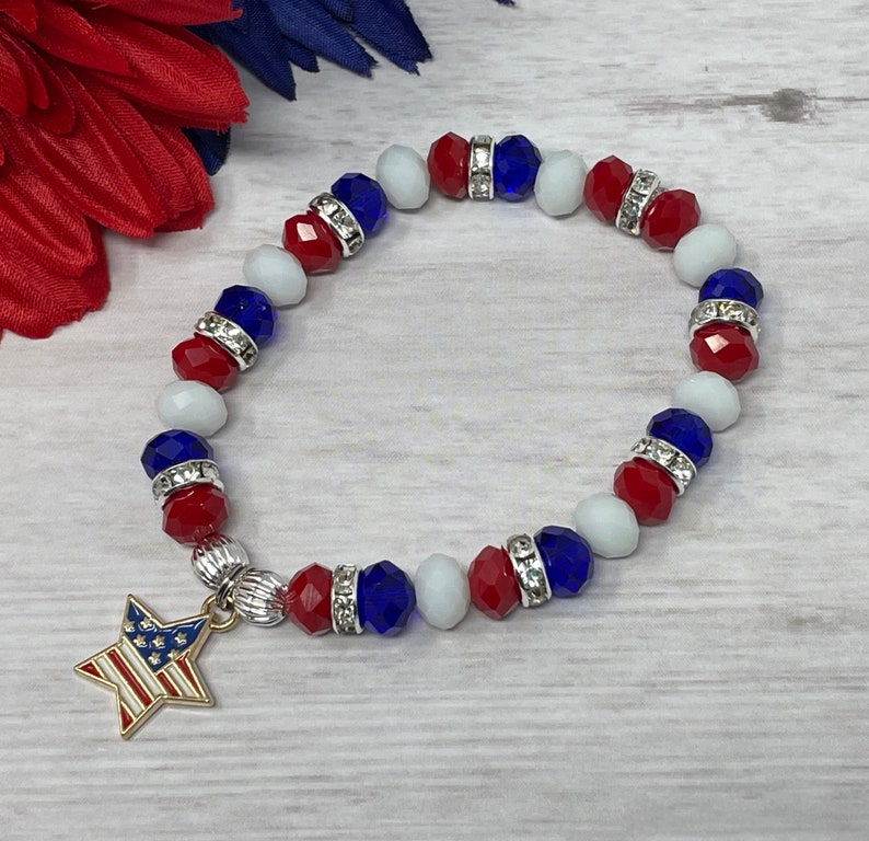 Pu&ograve; includere: Un braccialetto patriottico con perline rosse, bianche e blu, intervallate da distanziatori argentati. Un ciondolo a forma di stella dorata con un design a bandiera americana pende dal braccialetto. Il braccialetto &egrave; esposto su una superficie in legno chiaro.