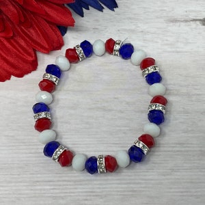 Puede incluir: Una pulsera de cuentas rojas, blancas y azules, con espaciadores plateados. La pulsera se exhibe sobre una superficie de vetas de madera clara, con detalles florales rojos y azules en el fondo.