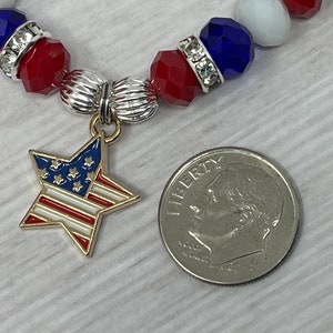 Pu&ograve; includere: Un braccialetto patriottico con perline rosse, bianche e blu, distanziatori color argento e un ciondolo a forma di stella con la bandiera americana. Un decimo di dollaro americano &egrave; accanto al braccialetto per il confronto delle dimensioni.