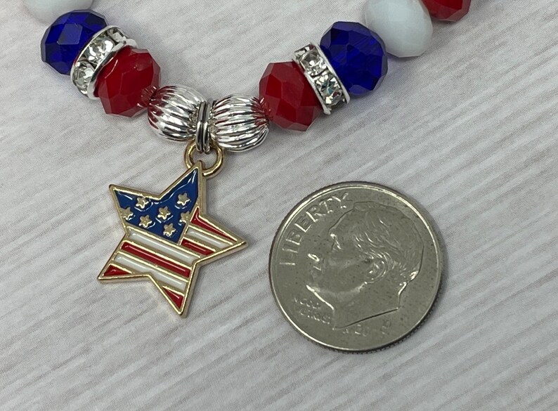 Pu&ograve; includere: Un braccialetto patriottico con perline rosse, bianche e blu, distanziatori color argento e un ciondolo a forma di stella con la bandiera americana. Un decimo di dollaro americano &egrave; accanto al braccialetto per il confronto delle dimensioni.