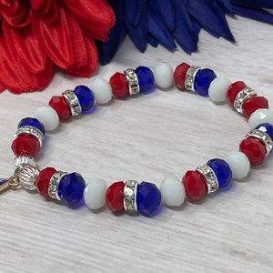 Pu&ograve; includere: Un braccialetto patriottico con perline rosse, bianche e blu, intervallate da distanziatori argentati. Un ciondolo a forma di stella dorata con un disegno della bandiera americana pende dal braccialetto. Il braccialetto &egrave; esposto su uno sfondo di accenti floreali rossi e blu.