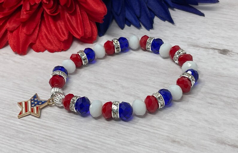 Pu&ograve; includere: Un braccialetto patriottico con perline rosse, bianche e blu, intervallate da distanziatori argentati. Un ciondolo a forma di stella dorata con un disegno della bandiera americana pende dal braccialetto. Il braccialetto &egrave; esposto su uno sfondo di accenti floreali rossi e blu.