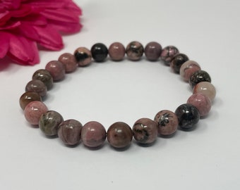 May include: A beaded bracelet featuring round, polished beads in shades of pink, brown, and black. The bracelet is made of natural stone beads, and is displayed on a white surface with a pink flower in the background. The bracelet is a piece of jewelry.