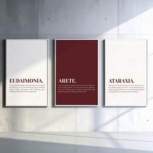 May include: Three rectangular wall art prints in thin white frames. The left print is white with the text EUDAIMONIA. The center print is deep red with the text ARETE. The right print is white with the text ATARAXIA. Each includes a definition.
