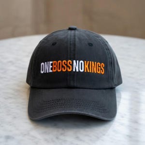 May include: A charcoal gray baseball cap featuring the embroidered text ONE BOSS NO KINGS on the front. The word ONE is in white, BOSS is in orange, NO is in white, and KINGS is in orange. The cap has a curved brim and a structured crown.