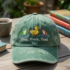 May include: A green baseball cap with an embroidered design featuring a frog, a duck, and a toad. The text "Frog, Duck, Toad" is embroidered below the characters. The cap is sitting on a wooden surface.